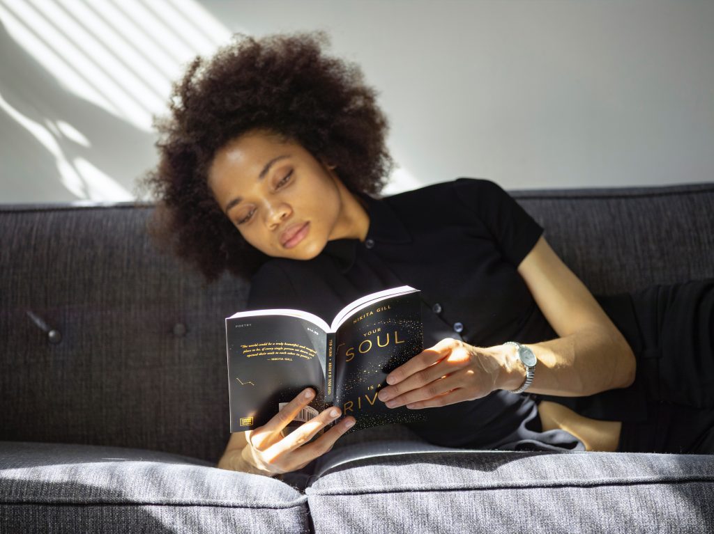Young Black woman lying on grey couch reading a book.