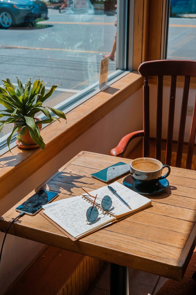 Open notebook with sunglasses on top, coffee and phone on a table beside a sunny window