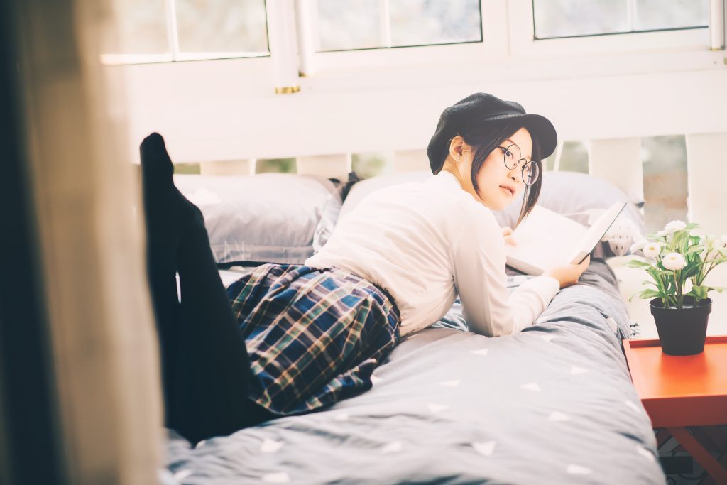 Asian woman with glasses and a page boy cap lying on her stomach on a bed reading a book.