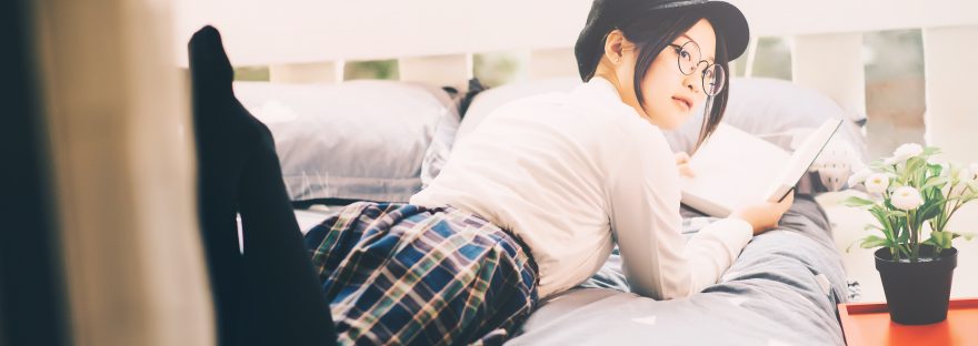 Asian woman with glasses and a page boy cap lying on her stomach on a bed reading a book.