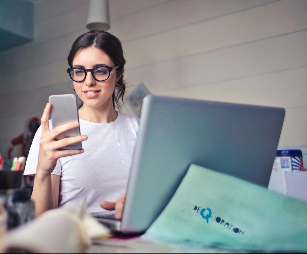 Young woman looking at phone in front of a sliver laptop.