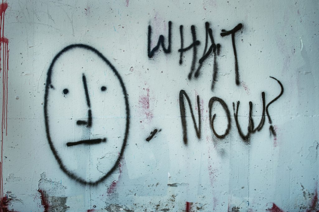 Grafitti of a face and the words "what now" on a white wall.