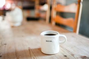 White mug filled with black coffee sitting on a wooden table with wooden chairs in the background. Mug says Begin.