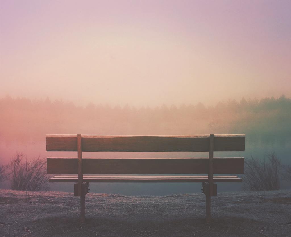 An empty wooden bench in the foreground with a misty pink view of trees and and a hazy purple sky.