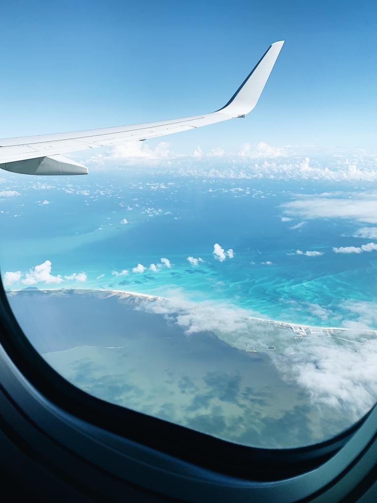 View through an airplane window of blue sky, blue water, white clouds, and the tip of an airplane wing.