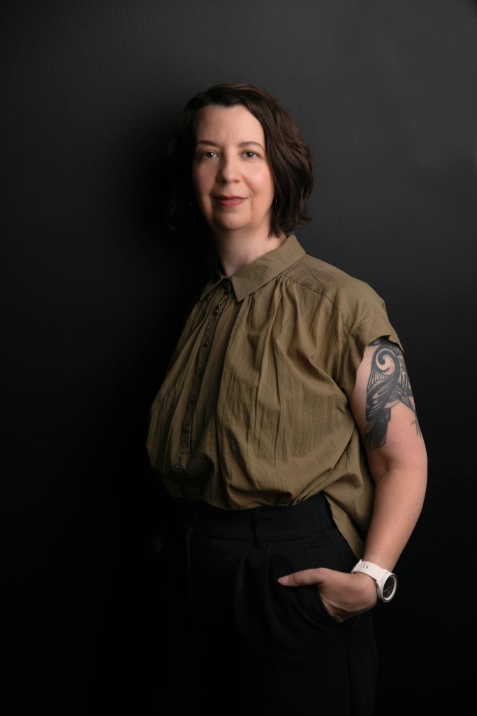 About me: Photo of Leslie Kern standing against a dark grey wall, wearing an olive green shirt and black pants.